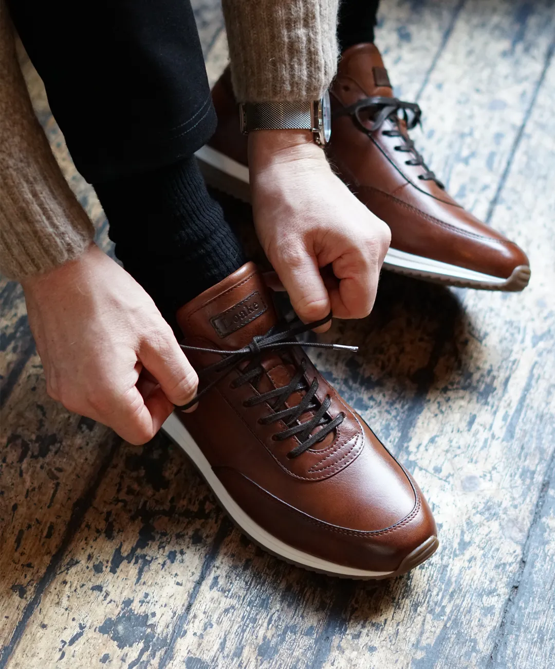 Man tying up his trainers in a public setting. Shoes shown are Bannister in cedar calf leather